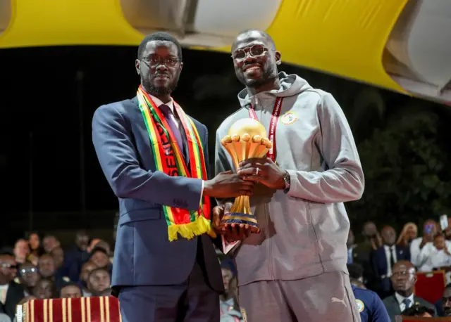 Le président sénégalais Bassirou Diomaye Faye et le capitaine de l'équipe nationale Khalidou Koulibaly célèbrent la victoire en Coupe d'Afrique des Nations en tenant ensemble le trophée.