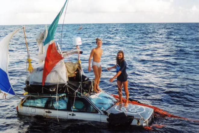 Deux hommes, Marco Amoretti et Marco De Candia, se tiennent debout sur le capot d'une voiture flottant sur l'océan. Des filets rouges, des cordes et des drapeaux sont attachés à la voiture. Ils ont les jambes nues, et l'un d'eux est entièrement nu, à l'exception de son caleçon blanc. L'autre porte un t-shirt bleu marine. Ils ont de longues barbes et des cheveux longs, et sont minces et bronzés.