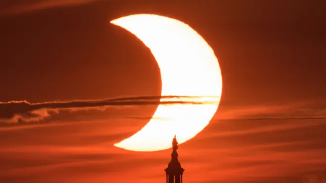 Lors d'une éclipse solaire partielle, le Soleil apparaît comme un croissant lumineux, avec la silhouette du sommet d'un bâtiment visible juste en dessous ; le ciel derrière lui arbore une lueur orange foncé.